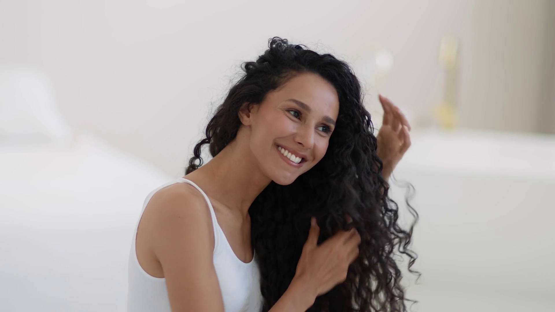 Smiling woman with long curly hair wearing a white tank top touches her hair in a bright room.